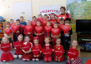Cała grupa ustawiona do zdjęcia wraz z nauczycielką na tle tablicy z napisem Walentynki. Dzieci trzymają czerwone poduszki i balony
